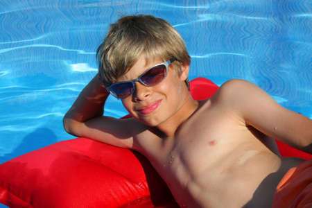 a 10-years old smiling American - German boy with sunglasses relaxing on an air mattress on the water surface of a swimming poolの写真素材