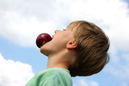 a handsome 7-years old boy making fun with a peach in his mouth, photographed in the summer sun with blue sky and clouds in the backgroundの写真素材