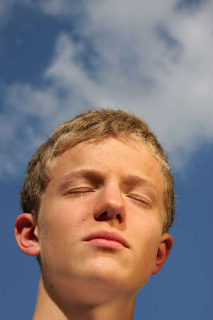 a handsome, smiling and happy 14-years old sun tanning teenager with a white shirt photographed in the summer sun with blue sky and clouds in the backgroundの写真素材