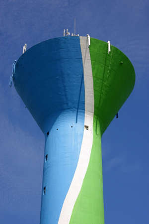 a colourful water tower, painted in blue, green and white, with cellular phone network antennas on top of it, photographed in the summer sun with blue and cloudy sky in the backgroundの写真素材