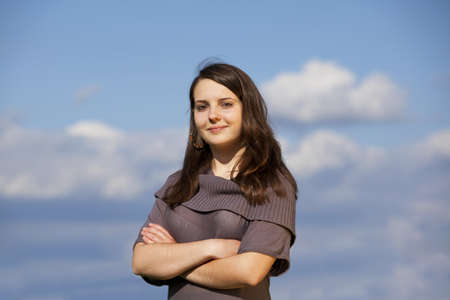 A beautiful and smiling teenage girl posing with her arms crossed, photographed with blue sky and clouds in the backgroundの写真素材