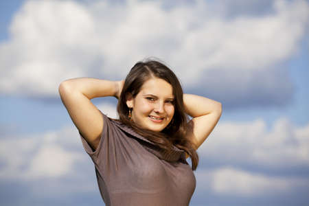 A beautiful and smiling teenage girl posing with her arms crossed behind her head, photographed with blue sky and clouds in the backgroundの写真素材