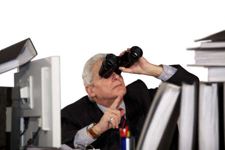 A senior businessman sitting behind his desk looking through binoculars and pointing with his finger