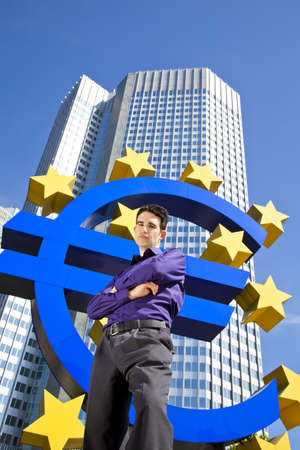 A cool looking businessman standing in front of the Euro symbol of the European Central Bank and posing with his arms crossedの写真素材