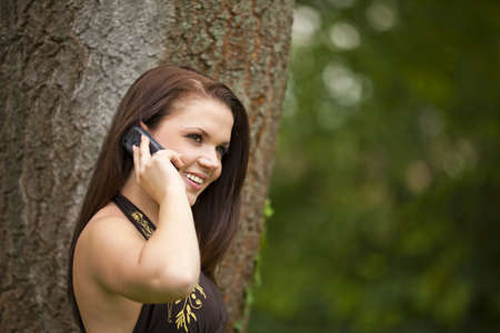 A smiling beautiful brunette woman in her twenties with a cell phone standing in a park in front of...