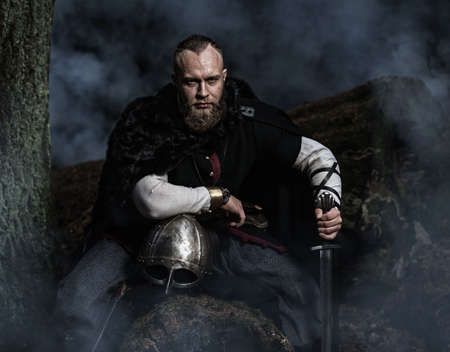 Viking with sword and helmet on a background of smoky forest. Warrior resting. Historic costume.