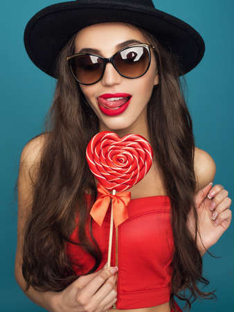 Love and valentines day, woman with sly smile holding heart. Portrait of Beautiful woman in hat with bright makeup and red heart in hand.の写真素材