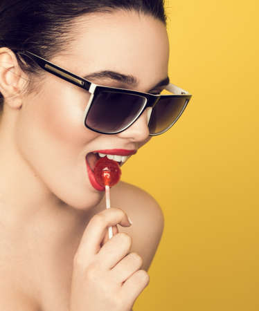 Beautiful amazing sexy smiling woman in sunglasses with a lollipop on yellow background. Brunette girl studio portrait licking candy.の写真素材