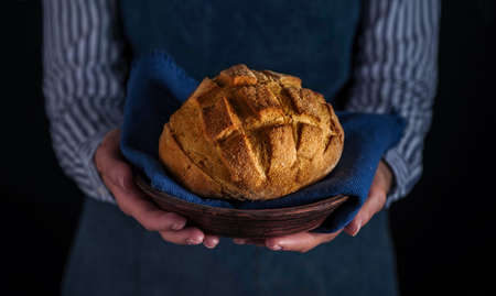 Baker's hands hold fresh bread over dark background.