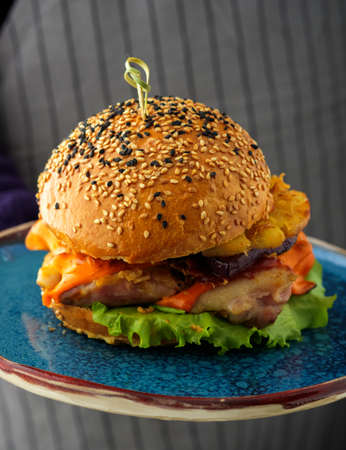 Tasty grilled chicken burger with lettuce, cheese, ananas and red onion served on blue plate. A waiter holding a plate with burgerの写真素材