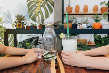 Two young adult women drinking sitting at a wooden table. Close up to glass and bottle, no face.の写真素材