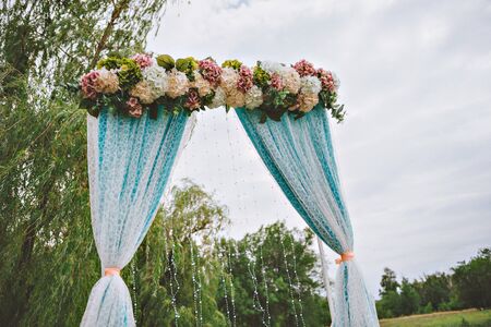 Beautiful white wedding arch decorated with purple and white flowers outdoors, near the willow treeの写真素材