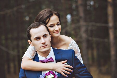 The bride hugs the groom. A pair of lovers in a pine forest. The groom sits on a log. The bride was behind me. Blue suit, white dress and serenity accessoriesの写真素材