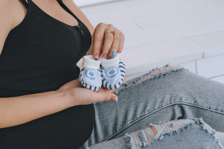 Happy pregnant woman holding booties, with a red bandage on his head. On a light background. Pregnancy red-haired young woman in a black t-shirtの写真素材