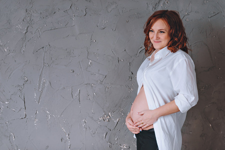 Pregnant woman in a white man's shirt, a studio portrait. The gray walls. Looks down. There is a place for text.の写真素材