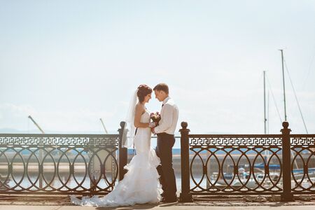 Smiling happy couple on bridge wedding day, the bride in a white dress with a train, a groom in a blue suit.の写真素材