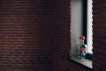 red flowers in a glass vase on the windowsill in the loft, brick aged wall. Background. Place for textの写真素材