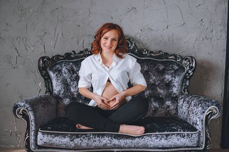 Young pregnant woman with red hair sitting on a gray sofa in the Baroque style. She's wearing a white shirt, visible to the naked pregnant belly. Looking at the cameraの写真素材