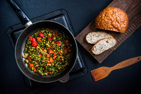 Vegetable stew with green peas, red pepper, onion and zucchini and spices. In a frying pan on a black background. On a wooden platform grain bread with flax seedsの写真素材