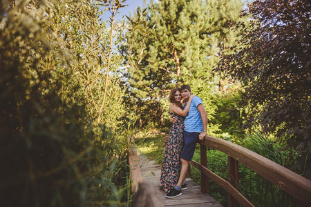 Happy romantic couple in the village, stroll on the wooden bridge. Young beautiful woman in long summer dress hugging the man. Look at the cameraの写真素材