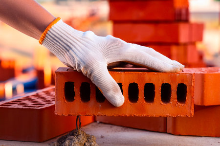 Gloved hand positioning perforated red brick during masonry work at construction siteの写真素材
