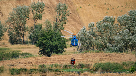 Firefighting helicopter flies with water bucket above wind turbines and forested hills