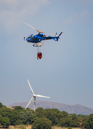 Firefighting helicopter flies with water bucket above wind turbines and forested hills