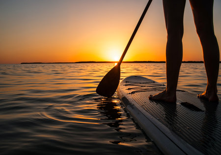Silhouette of paddleboarder on tranquil water during vibrant sunset, feet on the SUP boardの写真素材