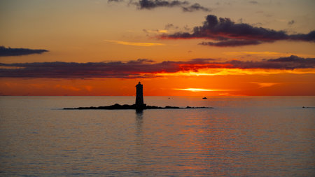 Peaceful lighthouse silhouette during vivid orange sunset over calm ocean watersの写真素材