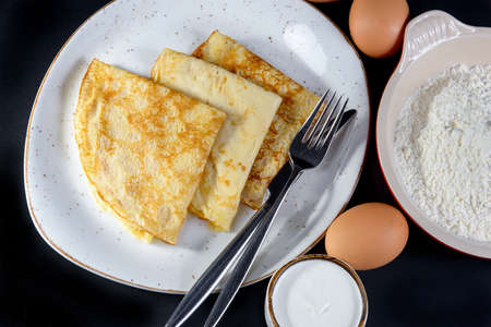 Pancakes on Shrove Tuesday with the ingredients on a plate on black background with eggs and flourの写真素材