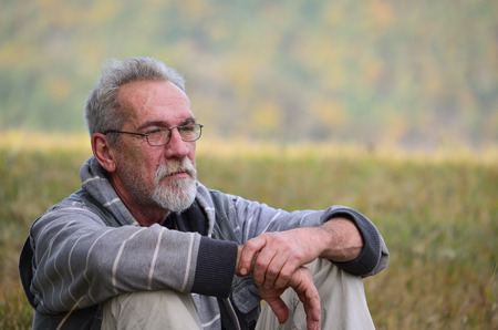Portrait of an elderly, worried  man with a gray beard and glasses sitting on grassの写真素材