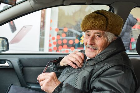 Vital elderly man with fur hat  in a car, smiling and holding glasses during drive