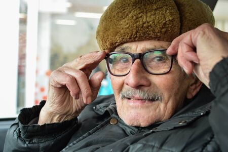 Portrait of active, vital elderly man with gray moustache and fur hat, sitting in a car and adjusting his spectaclesの写真素材