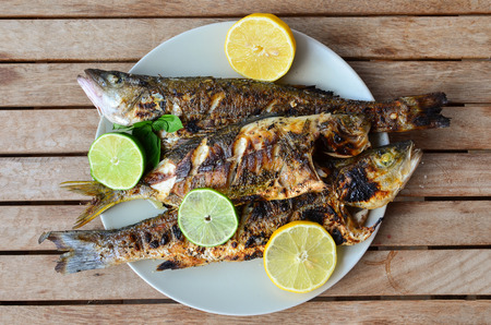 Fresh and healthy sea fish, seabass and salema porgy, grilled and served with lemon, lime and basil in a white porcelain plate on old wooden tableの写真素材