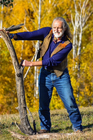 Strong, vital elderly man dressed in hippie style in outdoor exercise using big, heavy branches, on mountain meadow in front of autumn birch forestの写真素材