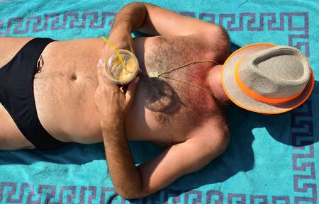 Beach joy concept, middle-aged man enjoys his coffee taking a nap in the sun on blue towel, with beach hat over his faceの写真素材