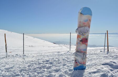 Snowboard left in snow vertically besides ski slope on the top of the mountain, in front of wooden fence, bright sunny winter day with clear sky but some low clouds in valley, lot of copy space, Kopaonik, Serbiaの写真素材