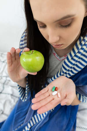 He holds a green apple in one hand and various pharmaceuticals in the other. Dressed in a bathrobe. A sick teenager.の写真素材