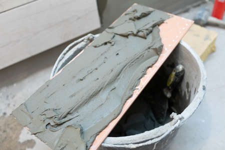 The bucket is on the platform right next to the legs of the worker who is laying the tiles.