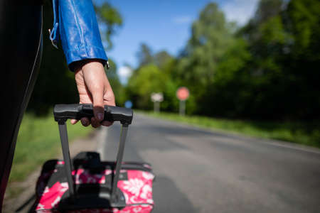 The teenagers hand gently clasps the handle of the travel suitcase. In the distance you can see an asphalt road and a green forest in full sun.の写真素材