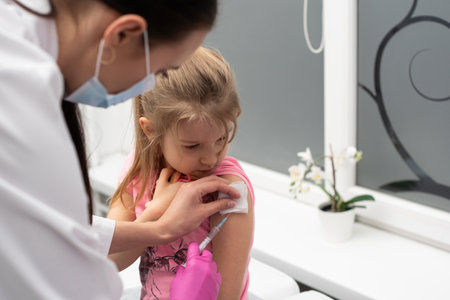 The nurse gently inserts the needle into the little girls arm. The child looks at the puncture site with the needle. A doctors office in a public hospital. Safe vaccination of children.の写真素材