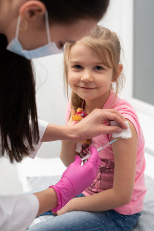 The girl looks into the lens while the doctor sticks the needle with the vaccine in her left arm. A doctors office in a public hospital. Safe vaccination of children.の写真素材