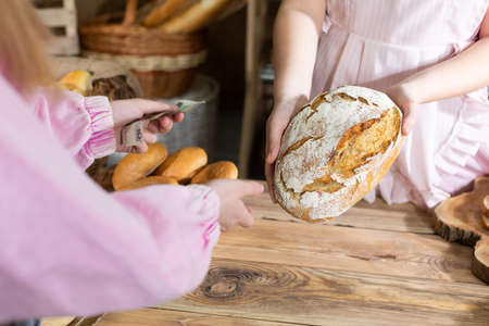 A teenager pays for selected rolls and bread in cash directly to the hands of a saleswoman from a...