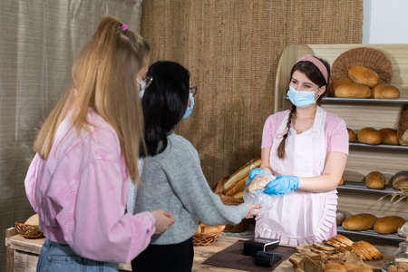 Two teenagers are waiting in line to buy bread from a bakery store. An attractive saleswoman serves...