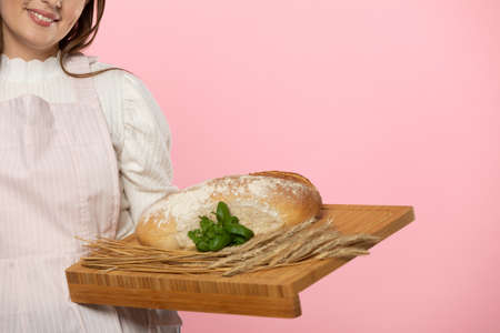 A young girl of beautiful European beauty holds in her hands on a wooden board a freshly baked loaf of bread decorated with green herbs and ears of grain. The whole composition is set against a uniform light pink background.の写真素材