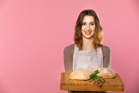 A young girl stands on a light pink background and in her hand she holds a board with two loaves of...