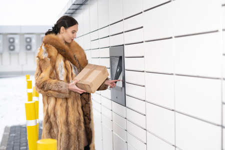 A young adult in a fur coat operates the touch panel of a parcel locker. Automatic machine for...