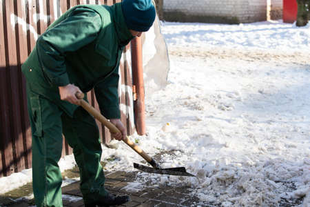 The landlord of the district in a housing estate in the city scrapes the snow off the pavement with a metal shovelの写真素材