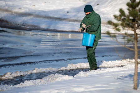 A manual worker walks with a blue bucket and sprinkles the pavements with expanded clay so that they are not slippery in the winter seasonの写真素材
