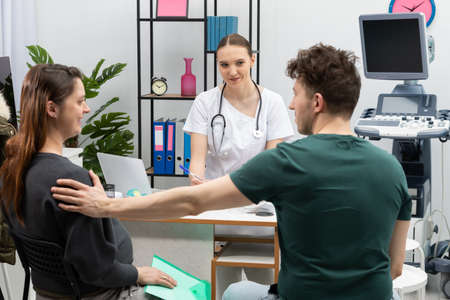 A married couple is sitting in an office with a gynecologist. The husband is supporting his pregnant...